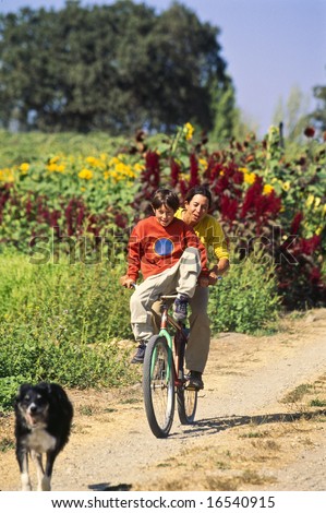 Older sister gives a ride to her younger brother on the handle bars of a bike as they inspect the family farm with their dog.