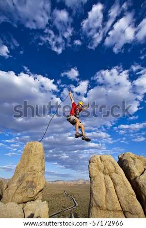 Male rock climber leaps across a gap on the summit of a pinnacle with a cloud filled sky behind him.