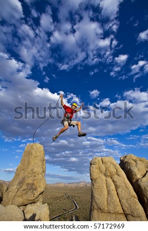 Male rock climber leaps across a gap on the summit of a pinnacle with a cloud filled sky behind him.