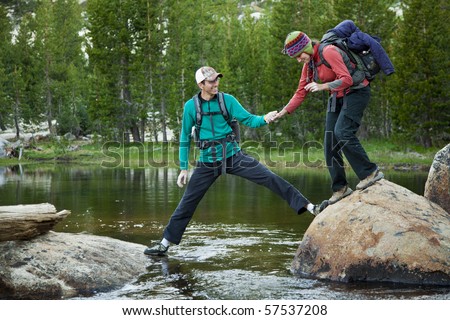 A young man grips the hand of his girlfriend as he helps her across a mountain stream.