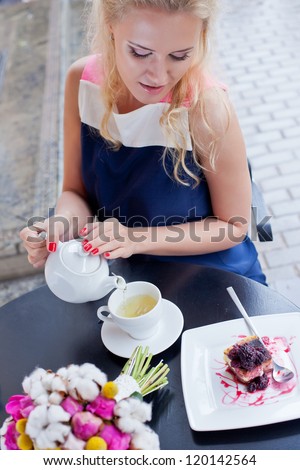 a beautiful young blond girl in summer dress at the table in pavement cafe is pouring green tea from the teapot into a white ceramic cup
