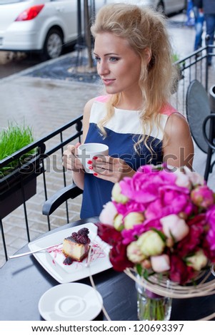 a beautiful young blond girl in summer dress at the table in pavement cafe holding a tea cup is looking thoughtfully in the distance