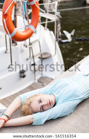 top view of beautiful blonde girl with blue eyes in transparent top lies on pier against white boat and life ring
