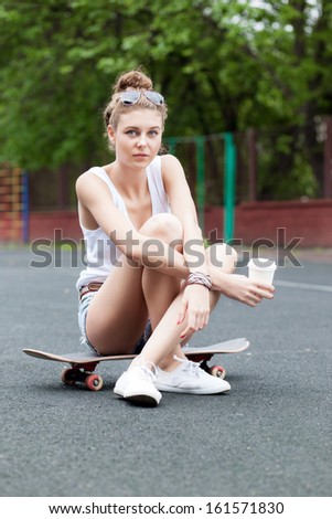beautiful young girl in jeans shorts sits on skateboard at basketball court holding a to-go cup