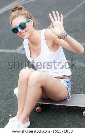 girl with curly drawn hair in white t-shirt and sunglasses with a white to-go cup sitting on skateboard at basketball court waves with her hand