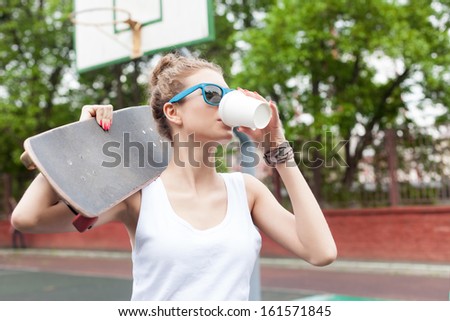beautiful girl in sunglasses drinks from white to-go cup holding skateboard on her  shoulder at basketball court