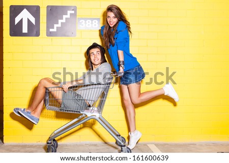 beautiful long haired girl in jeans mini skirt rides a boy in hat on shopping trolley in front of yellow brick wall