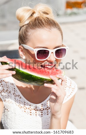 blonde girl with bow tie hair in white summer dress wearing sunglasses bites juicy watermelon