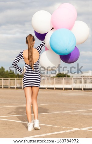 back view of girl in open-back black and white striped short dress and white high top sneakers holds a bunch of multicolored balloons