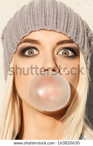Portrait Of Beautiful Young Blond Girl In Beanie Hat Blows Big Bubble From Bubble Gum