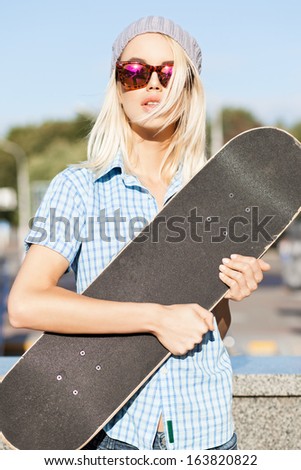 Close-up of beautiful  blond girl in beanie hat and leopard sunglasses who hugs skateboard on sunny day in the street