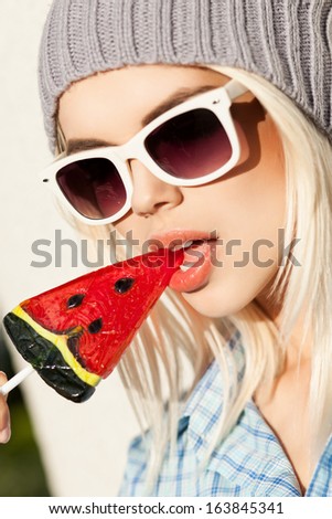 Portrait of sexy blond girl in white sunglasses and beanie hat who bites watermelon lollipop