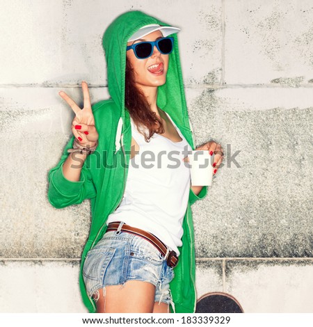 beautiful young girl in green hoodie and blue sunglasses with white to-go cup shows victory sign against grey wall