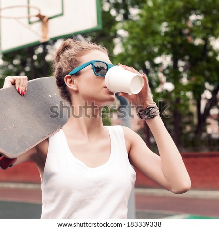 beautiful girl in sunglasses drinks from white to-go cup holding skateboard on her  shoulder at basketball court