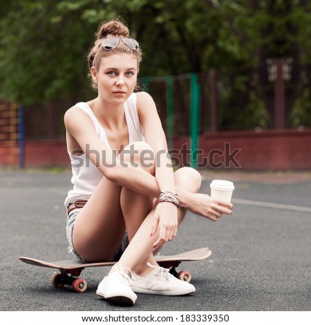 beautiful young girl in jeans shorts sits on skateboard at basketball court holding a to-go cup