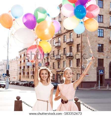 two smiling girls in white and pink dresses holding bunches of multicolored balloons in sun in the city street