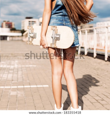 back view of young girl in short overalls with long legs and long silky hair holding skateboard