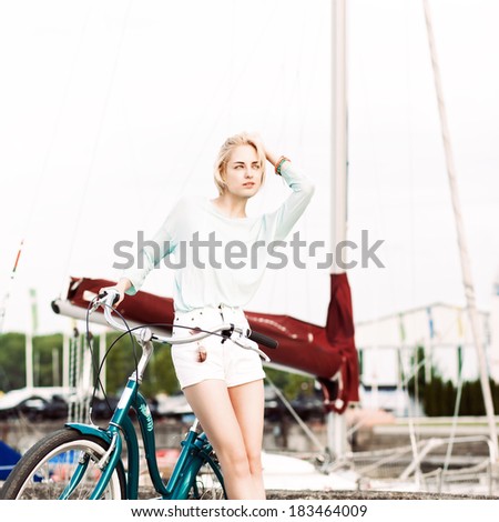 pretty tall blonde girl in short white shorts, transparent top with cruiser bike adjusts her hair at sea pier against yachts
