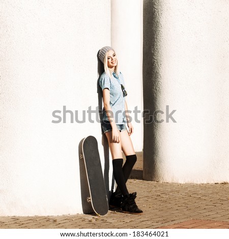 Happy smiling girl in short shorts and stockings with skateboard stands by columns on sunny day