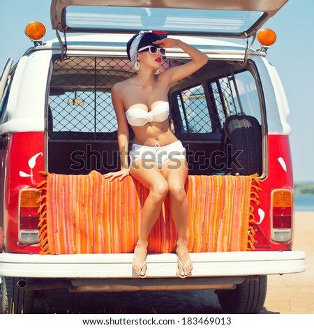 a beautiful young girl in retro look with red lips in a white swimsuit, a bandana and sunglasses is sitting in the car trunk and is looking in the distance