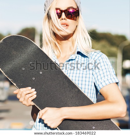 Close-up of beautiful  blond girl in beanie hat and leopard sunglasses who hugs skateboard on sunny day in the street