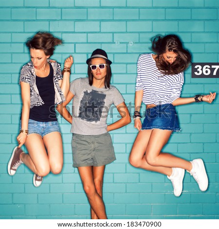 girls jump playing air invisible guitar while boy in hat and sunglasses poses for the camera in front of blue brick wall