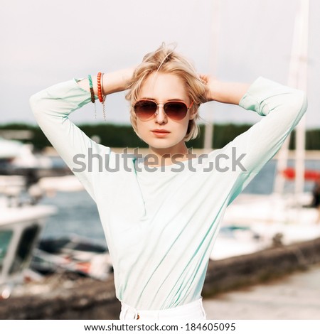 beautiful blonde girl in light summer style on city bike keeps her hands behind head at sea pier against yachts