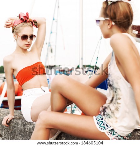 two beautiful girlfriends in fashion summer style sit against yachts