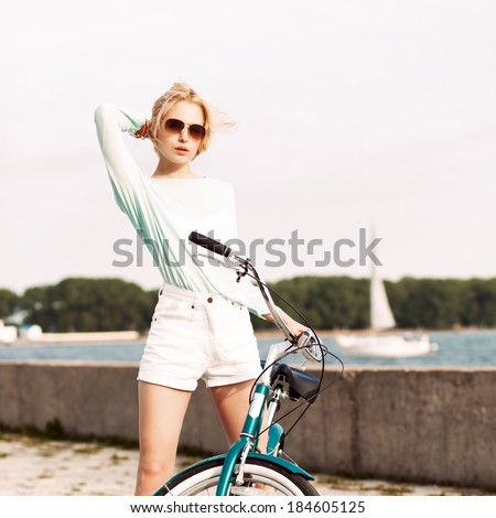 beautiful blonde girl in short white shorts, transparent top and sunglasses with cruiser bike at sea pier against yacht