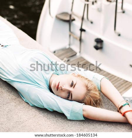top view of beautiful blonde girl with blue eyes in transparent top lies on pier against white boat and life ring