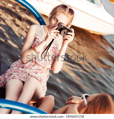 beautiful blonde girl in flower dress with photo camera takes picture of her tanned ginger girlfriend at sunset