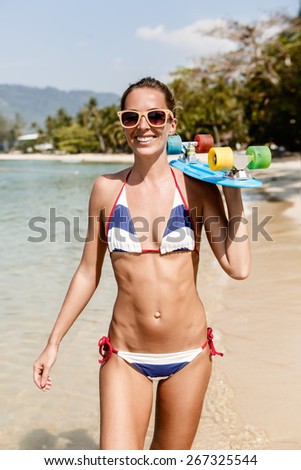 Beautiful sexy lady in blue and white striped bikini with penny longboard and sunglasses walks along the water line
