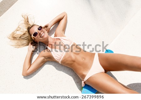 top view of sexy tanned blonde lady in sunglasses and bikini lying on blue penny board longboard in skate park