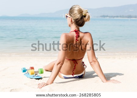 Beautiful sporty girl in blue and white striped bikini sits on sandy beach and sunbathing near her penny board
