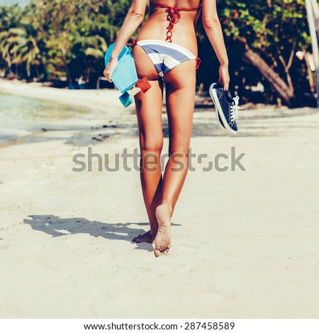 Blond sexy lady in blue and white striped bikini with penny long board and shoes in her hand walks along the water line