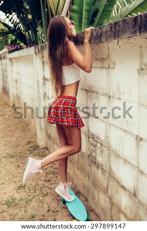 Sporty young lady with sexy butt in a red tartan mini skirt stands on blue penny skateboard trying to look over the fence of a tropical garden. Outdoor lifestyle picture on a sunny summer day.