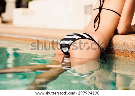 Close-up of athletic young beautiful lady in a black and white bikini clinging to side of the pool and raised on her hands on the edge of pool. Outdoor lifestyle picture on a hot sunny summer day.