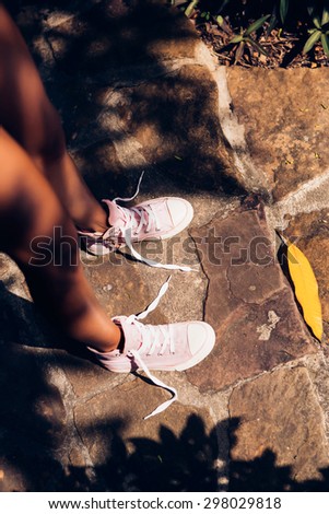 Beautiful athletic young woman in a yellow knitted bikini looks at her converse sneakers with untied shoelaces on the stone path in a tropical garden. Outdoor lifestyle on a hot sunny summer day.