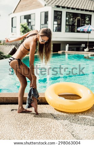 Athletic young girl with sporty ass in a striped bikini with yellow inflatable swimming ring takes off her jeans shorts at the refreshing pool. Outdoor lifestyle picture on a sunny summer day.
