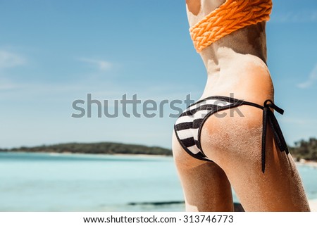Sandy buttocks of a young beautiful sporty lady in sexy striped black and white bikini panties on the tropical sea shore background. Outdoor lifestyle picture on a hot sunny summer day.