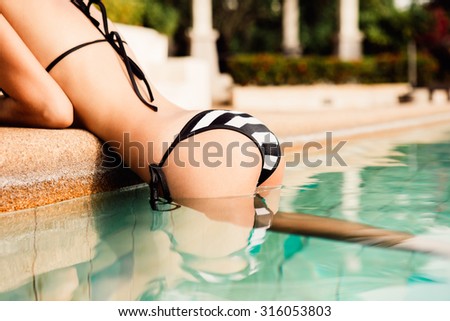 Closeup of a young beautiful sexy lady in a black and white bikini clinging to side of the pool and raised on her hands on the edge of pool. Outdoor lifestyle picture on a hot sunny summer day.