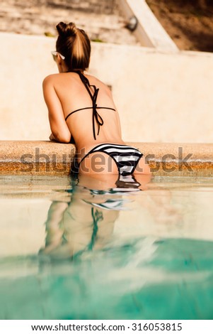 Close up of a skinny young beautiful girl in a black and white bikini clinging to side of the pool and raised on her hands on the edge of pool. Outdoor lifestyle picture on a hot sunny summer day.