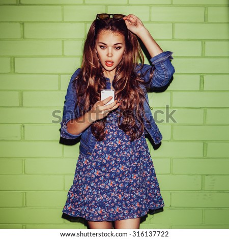 young happy beautiful long-haired brunette lady in blue dress with jeans jacket near the green brick wall with a surprised expression on her face looking to smartphone phone and correct her sunglasses