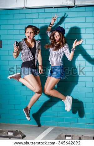 Lifestyle portrait of beautiful best friends hipster girls wearing stylish bright outfits and having great time. Jumping together in front of blue brick wall and have fun. Young active happy people.