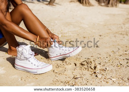 Outdoor lifestyle close-up of the legs of black beautiful teenage girl. Young hipster woman tying laces in her white sneakers. Sunny hot summer day at tropical seashore.
