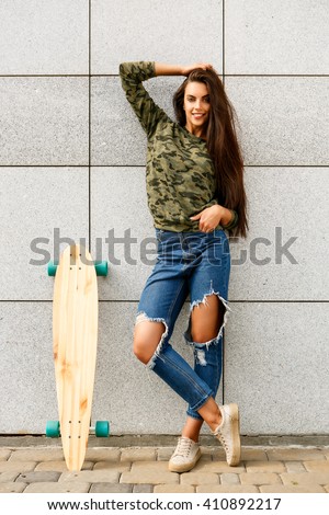 Portrait of happy beautiful woman with long sexy healthy hair in khaki top and ripped jeans with wooden longboard skateboard look to the camera and smile. Urban scene, city life. Hipster cute lady.