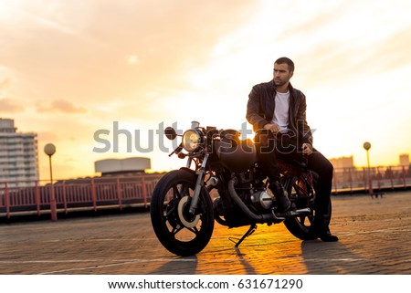 Handsome fit rider man with beard and mustache in black leather biker jacket sit on classic style cafe racer motorcycle at sunset time. Bike custom made in vintage garage. Brutal fun urban lifestyle.