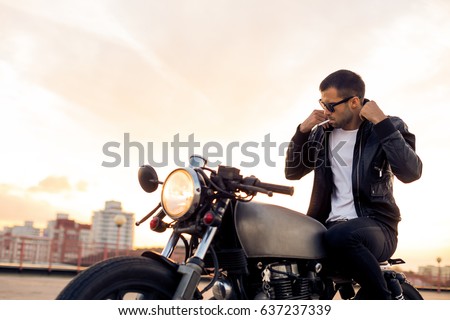 Handsome rider man with beard and mustache in black fashion sunglasses smoking cigaret and correct biker jacket sit on classic style cafe racer motorbike at sunset. Brutal fun urban lifestyle.