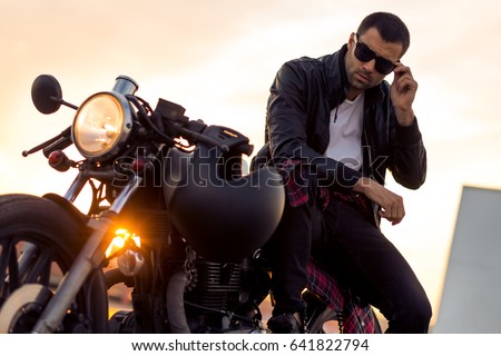Handsome rider guy with beard and mustache in black biker jacket take off sunglasses on classic style cafe racer motorcycle at sunset. Bike custom made in vintage garage. Brutal fun urban lifestyle.