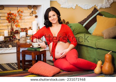 Happy pregnant woman in a red clothes sitting by the fireplace in the autumn interior